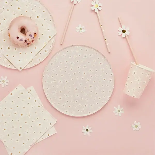 Daisy-themed party tableware with floral plates, napkins, cups, paper straws, and a donut on a soft pink background